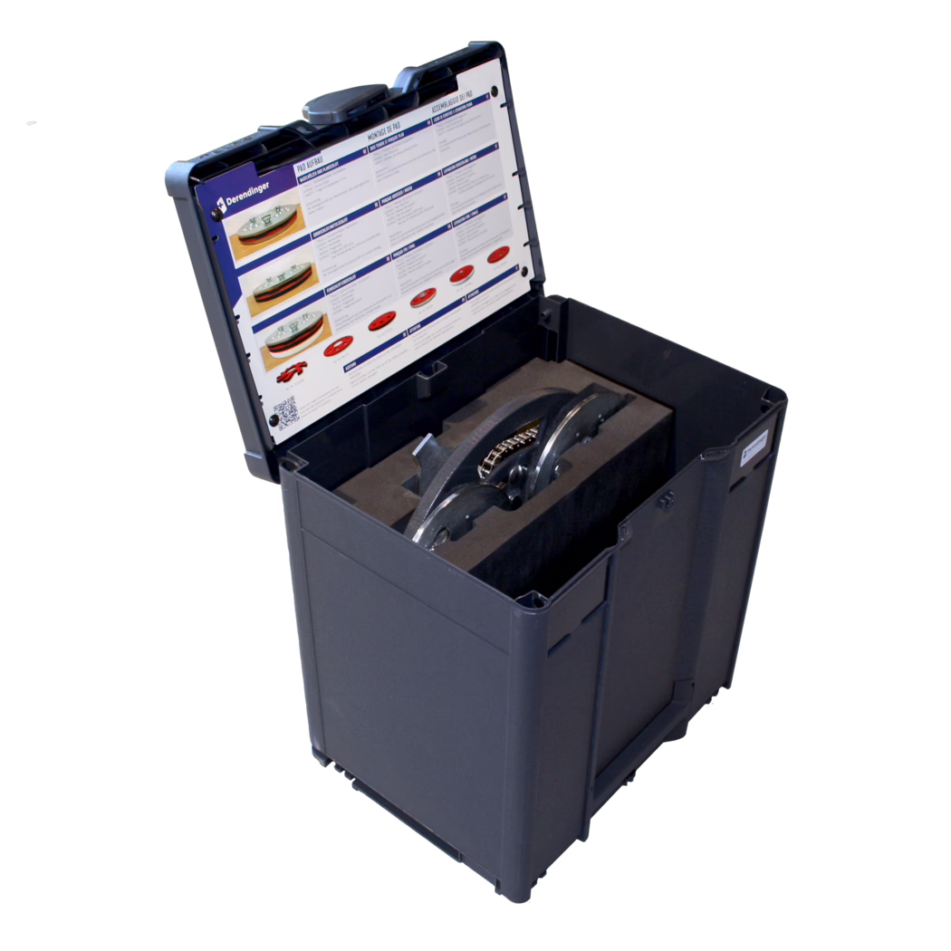 Derendinger_Dummy_178052_Dreischeibenteller_Box-DSW-400_Detail_2024.02 Mallette de transport en plastique noir ouverte avec des inserts en mousse découpés sur mesure qui maintiennent plusieurs composants métalliques lourds, notamment deux disques métalliques semi-circulaires reliés par une courte chaîne. Une feuille d'instructions et de montage avec images produit, icônes et colonnes pas à pas est fixée à l'intérieur du couvercle ; un petit logo « Derendinger » est visible en haut à gauche de la feuille. La mallette est photographiée en vue oblique depuis le dessus sur fond blanc, montrant clairement l'intérieur du couvercle et le compartiment doublé de mousse contenant les pièces métalliques.