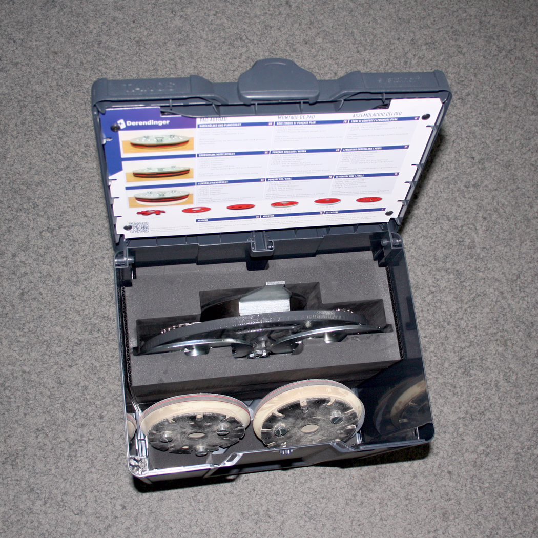 Derendinger_Dummy_178052_Dreischeibenteller_Box-DSW-400_Detail-2 Une vue détaillée d'une boîte à outils ouverte contenant un kit d'outils spécifique. Les outils comprennent un composant central avec une surface métallique brillante, entouré de rembourrage en mousse pour la protection. Deux disques circulaires sont visibles au fond de la boîte, chacun avec plusieurs trous et un bord en bois clair, ce qui suggère qu'ils sont des accessoires ou des pièces jointes pour l'outil principal. L'intérieur de la boîte présente une disposition structurée pour garder les outils organisés. Le haut de la boîte comporte un guide d'instructions imprimé, illustrant différentes options d'assemblage pour les outils, fournissant des instructions visuelles en plus d'un texte écrit.