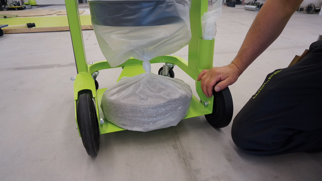 WOLFF_Application_Image_VacucleanLP_3 Un gros plan d'un chariot vert avec un sac en plastique rempli de matériel en bas. Le chariot a deux roues noires visibles au sol, et la main d'une personne est vue en train d'ajuster le chariot ou son contenu. L'arrière-plan présente un espace de travail avec des outils et des matériaux éparpillés, indiquant un projet en cours.