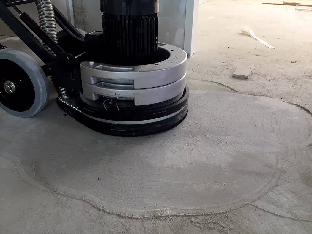 WOLFF_Application_Image_Neo400_1 Gros plan d'une ponceuse de sol en train d'uniformiser une surface en béton sur un chantier ; on distingue le carter du moteur, la tête de ponçage circulaire et les roues, ainsi que des traces de ponçage et de la poussière sur le sol en béton gris sous un éclairage intérieur froid. Aucune personne visible.