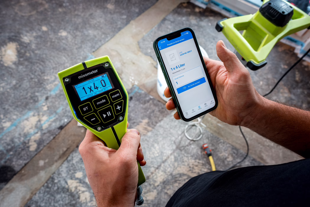 Deux mains tiennent un appareil portable vert vif appelé « mixometer » et un smartphone affichant une application de mélange. L'écran rétroéclairé du mixometer indique « 1 x 4.0 » et le code produit « UZIN NC 585 F » ; l'écran du téléphone montre une fiche produit avec « 1 x 4 Liter » et un bouton bleu « An Gerät senden ». La scène se déroule sur un sol de chantier ou de rénovation avec des câbles épars, un pied/outil jaune‑vert et un tournevis posé en arrière‑plan. L'image met l'accent sur les appareils et les mains de l'utilisateur, illustrant le contrôle numérique du dosage des matériaux sur un chantier de rénovation.