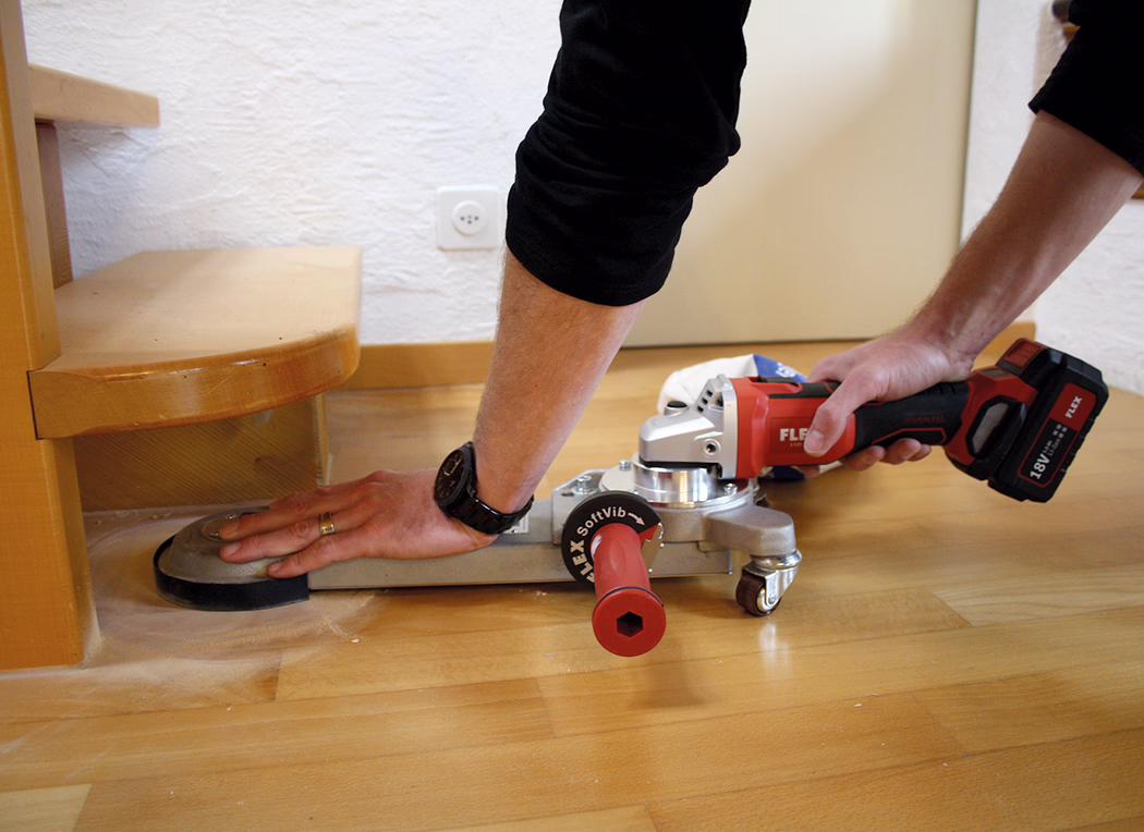 Derendinger_Application_Image_URM-DS_Akku_2 Close-up of a person sanding a wooden floor next to a stair step using a red battery-powered floor sander attachment connected to a FLEX angle grinder. The image shows the person's forearms and hands: the right hand grips the grinder's handle while the left hand steadies the flat sanding plate sliding along the floor edge. A small caster wheel supports the metal adapter, and fine wood dust is scattered on the floor. A wristwatch and a wedding ring are visible on the person's left hand; a white wall socket and a wooden baseboard are visible in the background. The scene suggests indoor floor renovation work carried out carefully along the step edge.