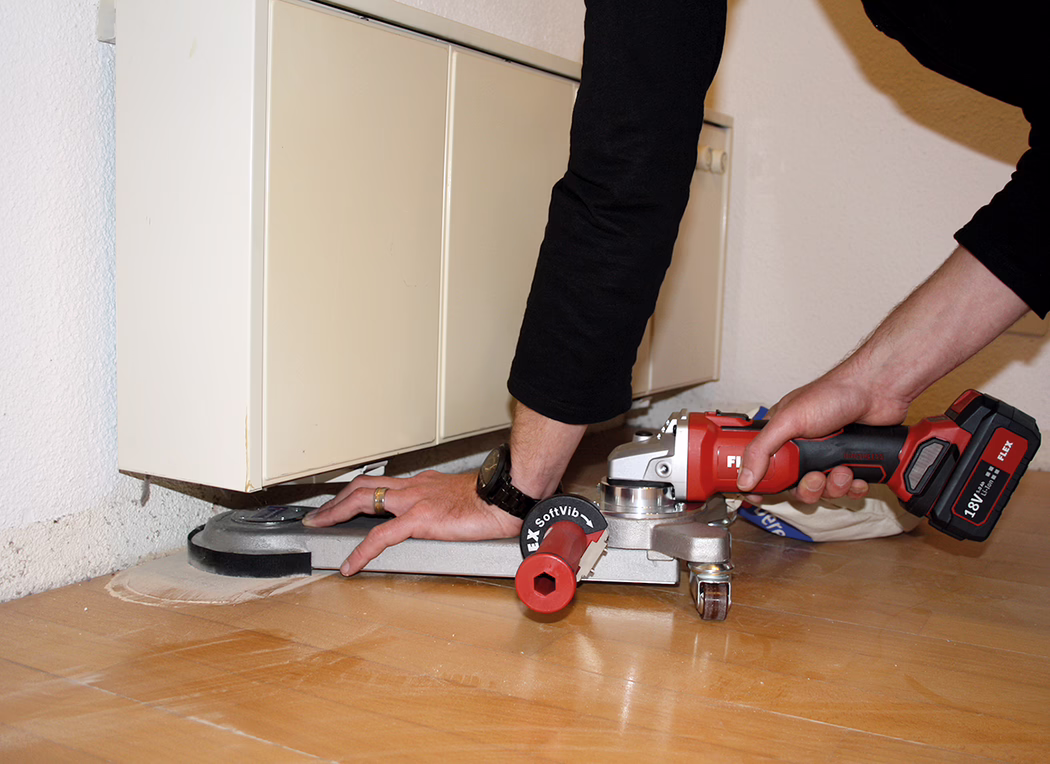Derendinger_Application_Image_URM-DS_Akku_1 Gros plan d'une personne utilisant une meuleuse sans fil rouge fixée à une base de ponçage pour poncer ou décaper un parquet en bois sous le coffrage d'un radiateur mural. Seules les mains et les avant-bras sont visibles ; la personne porte un vêtement noir à manches longues et une montre. L'outil est marqué 'FLEX' et possède une batterie 18V.