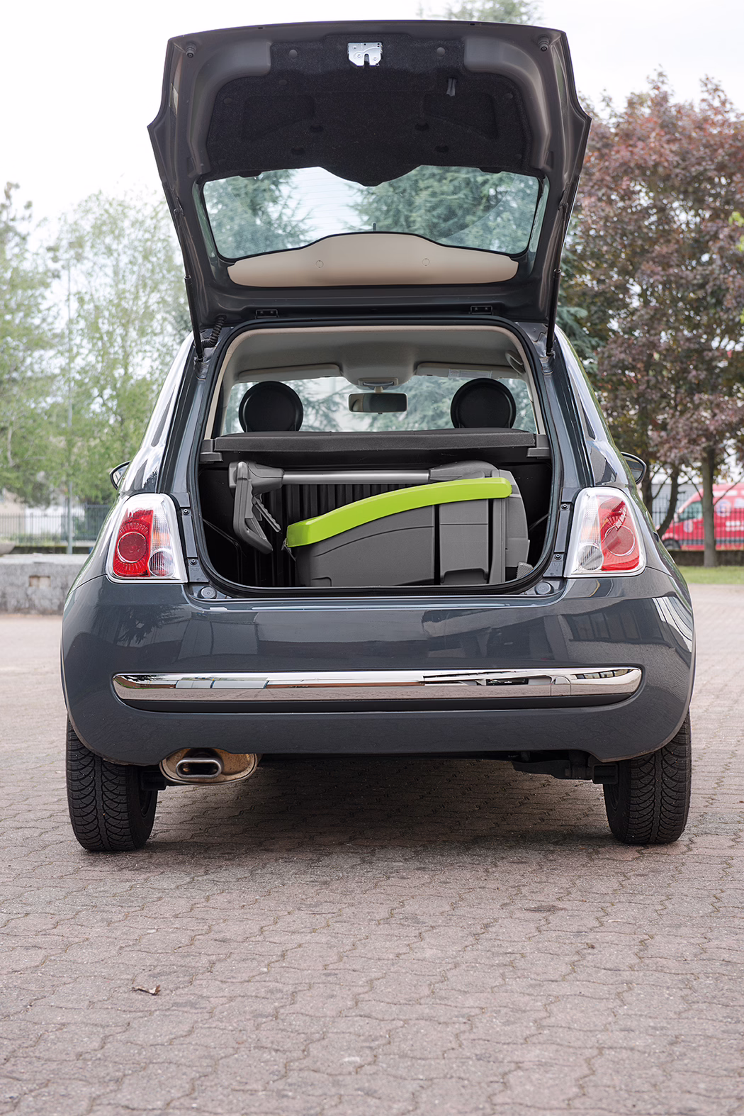 WOLFF_Product_detail_Cleanfox_Kofferraum_2017 Vue arrière d'une petite voiture compacte gris foncé avec son coffre ouvert ; un siège enfant plié ou rehausseur avec une poignée vert clair est posé dans le coffre ; stationnée à l'extérieur avec des arbres et du pavage en arrière-plan.