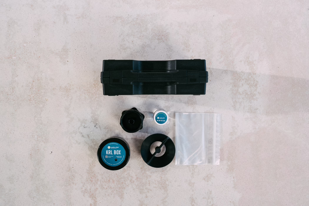 WOLFF_Campaign Images_KRL Box_2025-08 (44) Top-down view of a light concrete surface with a small black plastic toolbox centered near the top and several round and cylindrical components arranged below. Visible items include a round container labeled "KRL BOX" with a blue circular label, a smaller jar with similar blue branding, a black funnel-shaped part, a ring-shaped black plastic piece, and a transparent resealable plastic bag. The scene is minimal and neatly organized, photographed from above with soft natural lighting and the textured concrete background clearly visible.