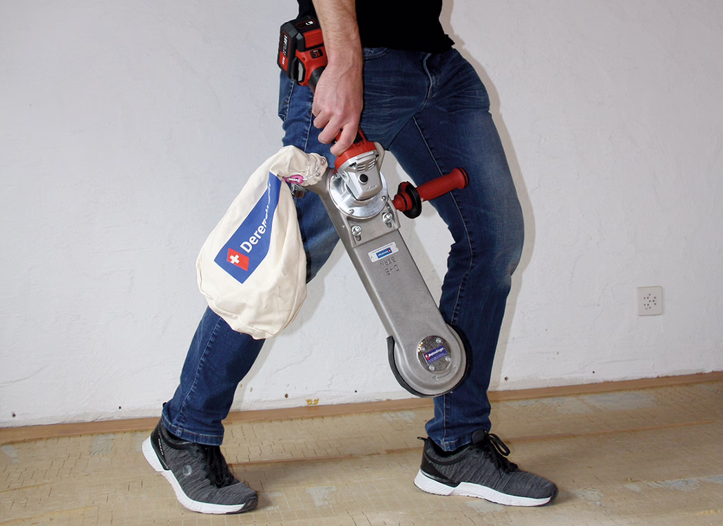Derendinger_Application_Image_URM-DS_Akku_3 Une personne, visible seulement de la taille aux pieds, porte un jean bleu et des baskets noires et tient une ponceuse de sol portative avec un sac à poussière. L'outil a une poignée rouge et un boîtier métallique ; le sac en toile présente un logo bleu avec une croix blanche. On voit un sol en bois et un mur blanc.