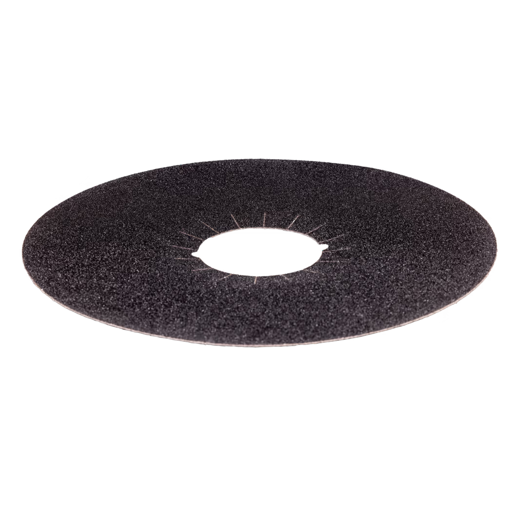 Close-up photograph of a circular abrasive grinding disc placed on a white background. The disc surface is covered with coarse black grit and shows a round central hole with radial slits around the inner edge; only the upper portion of the disc is visible in the frame. The image highlights the rough, granular texture and individual abrasive particles, with even bright lighting that emphasizes surface details and slight wear marks near the center.