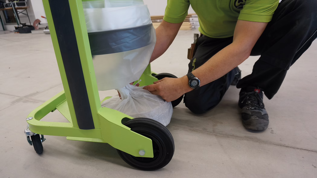 WOLFF_Application_Image_VacucleanLP_2 Une personne à genoux à côté d'un chariot vert avec des roues. Le chariot a un sac poubelle dans un support, et la personne est en train de nouer le sac poubelle. L'environnement est un atelier avec des outils visibles en arrière-plan.