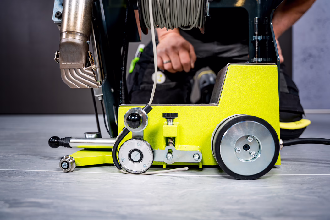 Gros plan d'une machine industrielle de sol de couleur jaune vif avec des roues, une buse métallique, un câble enroulé sur un tambour et la main d'un ouvrier visible en arrière‑plan ; l'image met en valeur des détails mécaniques et des surfaces texturées.