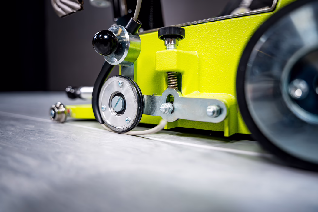 Gros plan d'un appareil mécanique vert citron sur une surface métallique, montrant une petite roue guide, des pièces métalliques, un ressort visible, des vis, un bouton de réglage noir et un câble connecté.
