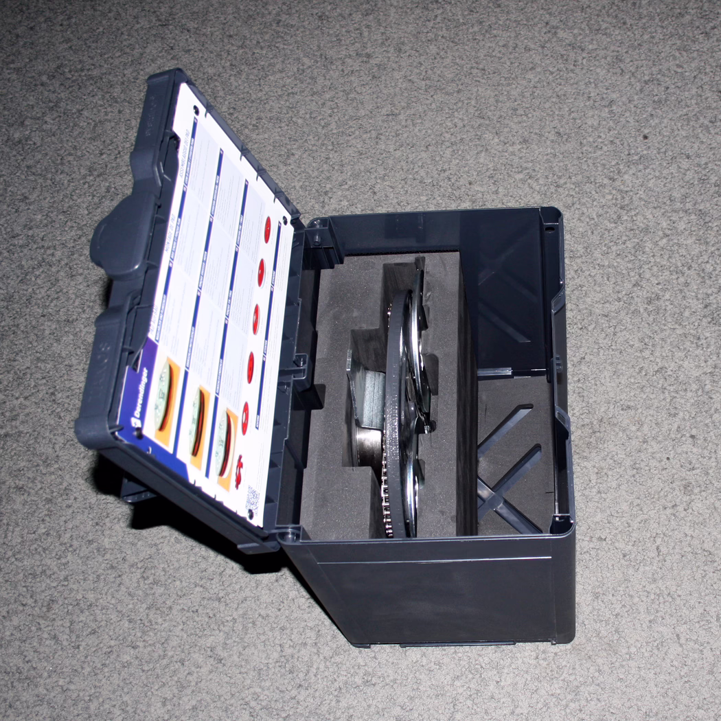 Derendinger_Dummy_178052_Dreischeibenteller_Box-DSW-400_Detail_2023.12 Une vue rapprochée d'une boîte à outils de couleur sombre ouverte pour révéler son intérieur. La boîte à outils contient un composant métallique circulaire, probablement un engrenage ou une partie d'un assemblage mécanique. Au-dessus du composant se trouve une feuille d'information imprimée avec divers icônes et textes, indiquant probablement différents réglages ou utilisations pour le contenu de la boîte. L'intérieur de la boîte à outils a un insert en mousse pour la protection, tandis que l'extérieur semble robuste et durable. La surface sous la boîte à outils est un tapis gris uni, offrant un fond neutre.