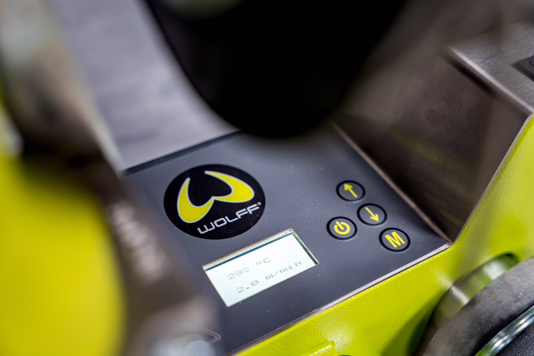 Gros plan d'un panneau de commande numérique ou d'un instrument de mesure monté sur une machine jaune-vert fluorescent de la marque 'WOLFF'. L'image met en avant un logo rond noir et jaune WOLFF, un petit écran LCD rétroéclairé affichant des valeurs de mesure, et plusieurs boutons ronds noirs avec des symboles jaunes (marche/arrêt, flèches, 'M'). Le boîtier métallique environnant est peint en jaune-vert vif et une surface en acier inoxydable brossé est visible en haut à droite. Une faible profondeur de champ rend le logo et les boutons nets tandis que les bords et l'arrière-plan sont légèrement flous, soulignant l'interface et la texture industrielle de l'appareil.