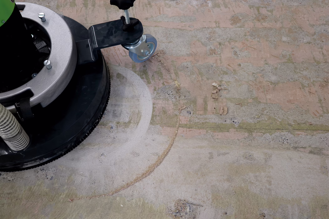 WOLFF_Application_Image_Neo400_2 Close-up of a floor polishing machine: visible are a circular black polishing pad, part of a metal housing and a small swivel caster wheel, positioned on a worn concrete or adhesive-covered floor. The floor surface shows a semicircular sanding mark made by the machine, patches of greenish residue or mold, torn adhesive fragments and scattered small debris, revealing the rough substrate beneath; a corrugated vacuum hose is visible at the left edge.